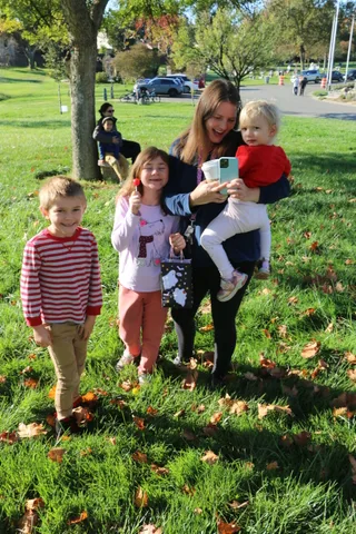 A mother holds two young children close while a third child stands nearby, all smiling.