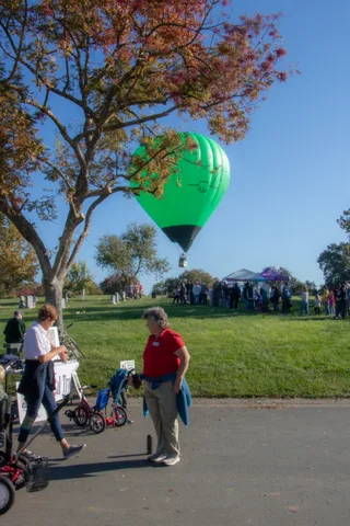 A small green hot air balloon floats over the park as scattered guests watch near a tent.