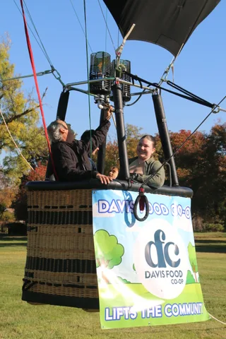Two riders peek over the edge of the balloon basket above the sponsor banner.