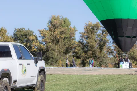 A white support truck is parked in the grass with the balloon basket small in the distance.