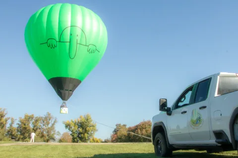 A small green hot air balloon floats above the park with a support truck below.
