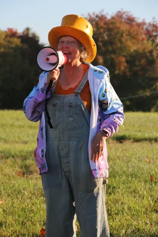 A woman in an orange top hat speaks into a megaphone with a wide smile.