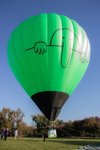 A second view of the green hot air balloon's cartoon-face envelope against blue sky.