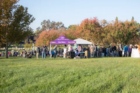 A crowd gathers near a canopy tent across a wide grass field lined with autumn trees.
