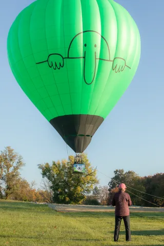 The balloon's dark canopy descends close to the treeline as one person steadies the line below.