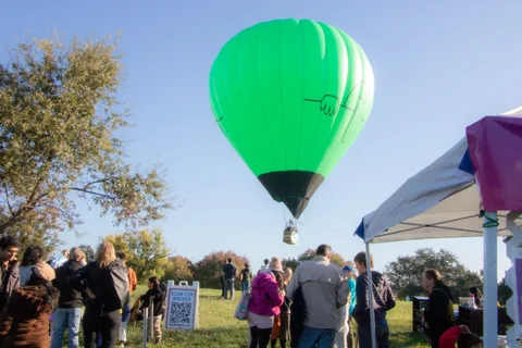 The green balloon floats low over a crowd of visitors near the event tents.