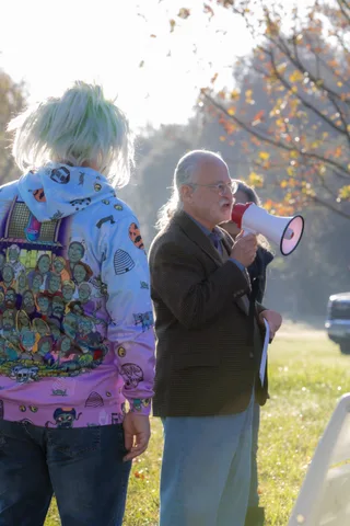 A person in a wild patterned wig and costume stands beside a man holding a megaphone.