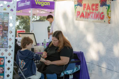 A volunteer paints a boy's face at a booth advertising face painting and vegan snacks.