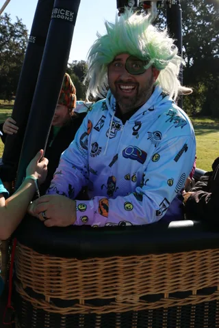 A man in a green wig and sunglasses grins in a colorful festival jacket.