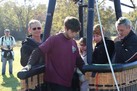 A small group stands together beside a hot air balloon basket on the grass.