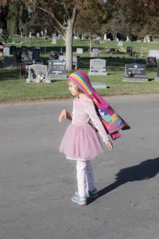 A young girl in a pink tutu and colorful wrap walks along a cemetery path.