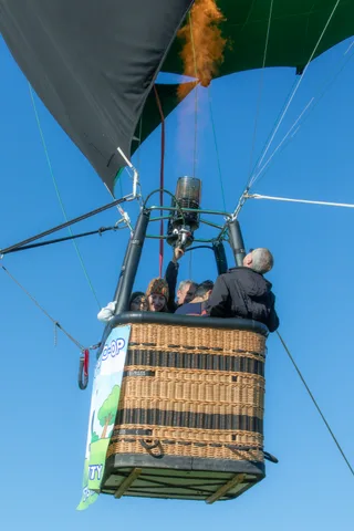 Two riders stand at the edge of the balloon basket, a red rope crossing the sky behind them.