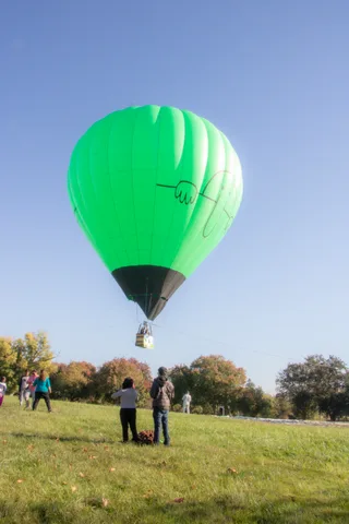 A dark hot air balloon rises over the grass field as two people watch, golden trees on the horizon.