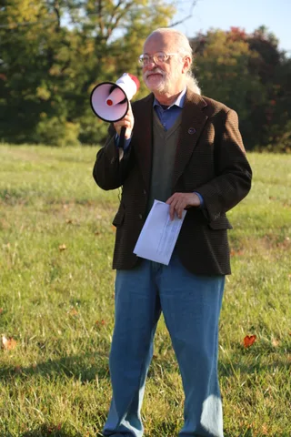 A man in glasses and a blazer speaks to the gathered crowd.