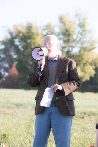 An older man raises a red megaphone to speak, backlit by warm morning light.