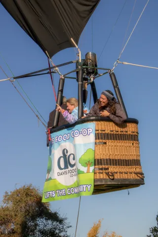 A child in a knit cap waves from a balloon basket beside a laughing woman against blue sky.