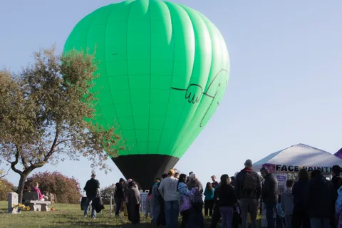 A large green hot air balloon looms low over a crowd gathered on the cemetery lawn.