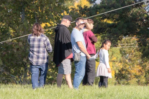 A family stands together watching a hot air balloon rise over the autumn trees.