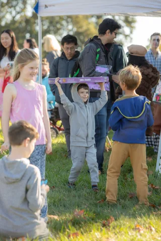 A man and boy hold up a decorated box together as a girl in pink walks past.