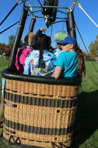 A costumed rider in a green wig reaches up inside the balloon basket with another passenger.