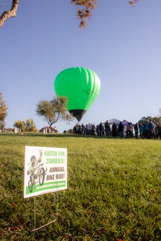 The balloon rises above a distant line of people in the field, a Zombie Bike Ride sign in the foreground.