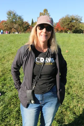 A woman in sunglasses smiles on the grass with autumn-colored trees behind her.