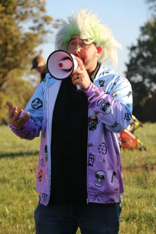An emcee in a green wig raises his arm mid-announcement on the lawn.
