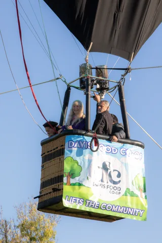 Riders lean over the edge of a hot air balloon basket wrapped in a Davis Food Co-op banner.