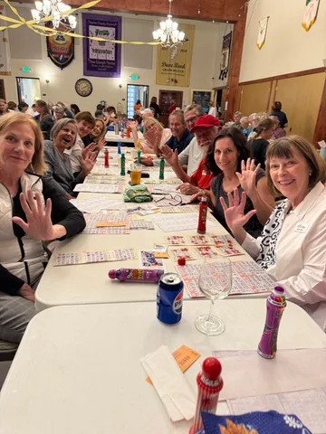 A long table of bingo players waves at the camera, daubers and bingo sheets spread out in front of them.