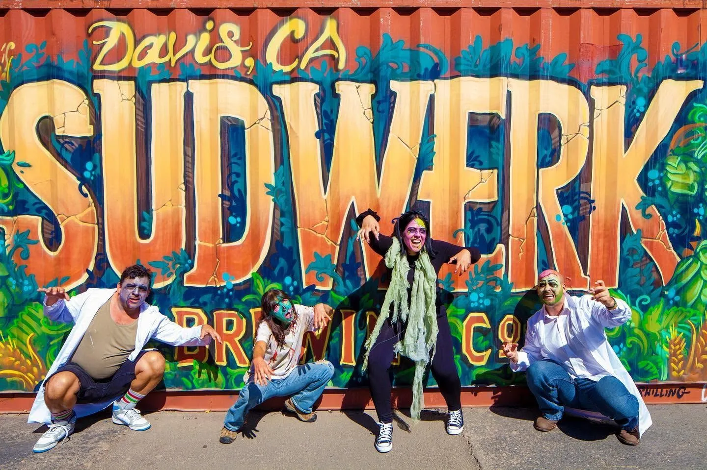 Four people in zombie make-up crouch and pose in front of a large painted Sudwerk Brewing Co mural reading Davis, CA.