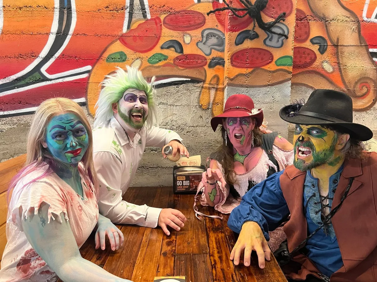 Four people in green zombie make-up lean across a table in front of a painted pizza mural at Woodstock's.