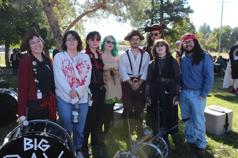 A band of eight in pirate and gothic costumes poses together behind their bass drum reading Big Day.