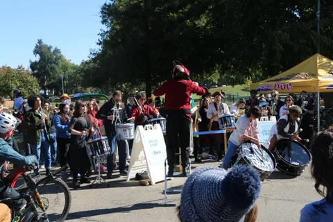 A costumed marching band plays drums and horns for a crowd gathered in the park.
