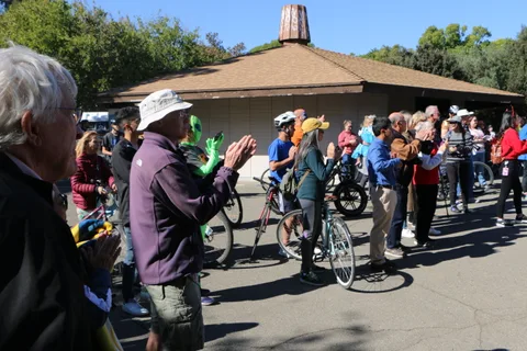 A crowd of cyclists applauds beside a park building on a sunny day.
