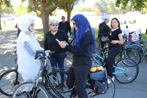 Costumed cyclists in wigs and face paint chat beside their bikes under park trees.