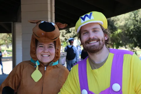 A couple dressed as Scooby-Doo and Wario grin for the camera under a covered pavilion.
