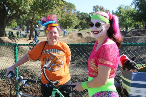 Two costumed cyclists in colorful wigs pause together on the bike path.