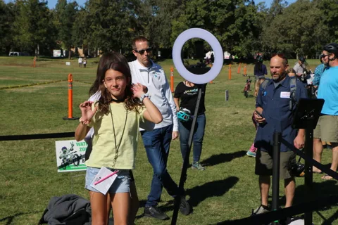 A girl in a yellow shirt dances past a ring light near a festival sign.
