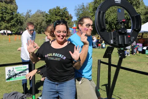 A woman with zombie makeup waves playfully for a festival photo booth camera.