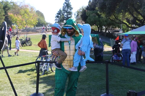 A man in a dinosaur costume carries two children dressed as a superhero and a shark at the festival.