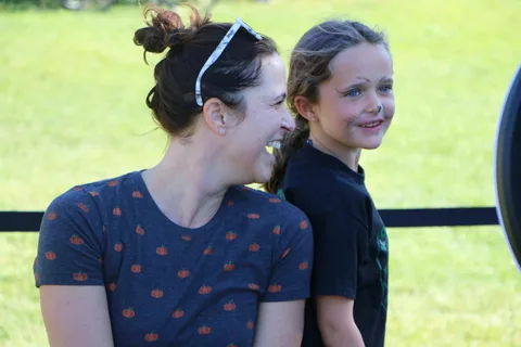 A mother and her cat-painted daughter laugh together on a sunny lawn.