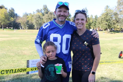 A man and woman smile with a child holding a shaved ice cup on the park lawn.