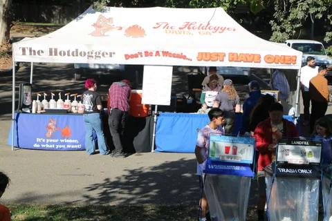 A hot dog vendor tent labeled The Hotdogger serves festivalgoers with condiments lined up on the table.