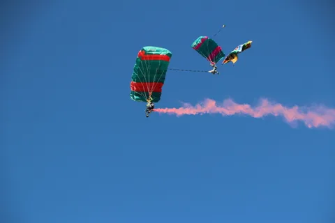 A skydiver trails pink smoke beneath an open parachute against a clear blue sky.