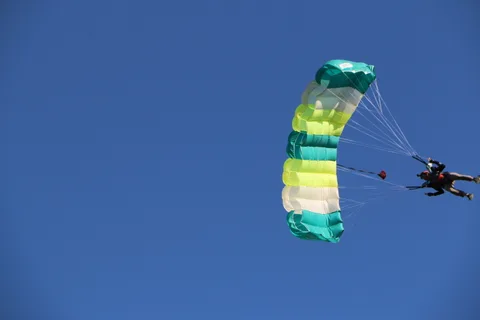 A skydiver descends beneath a striped teal and yellow parachute against clear blue sky.