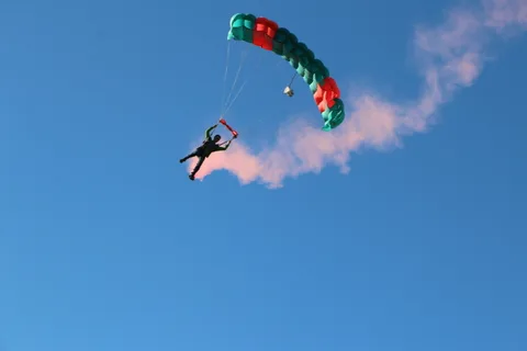 A skydiver descends with a trail of orange smoke against an open sky.