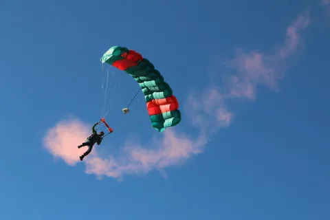 A skydiver reaches for a red toggle beneath a red and teal parachute with smoke drifting below.