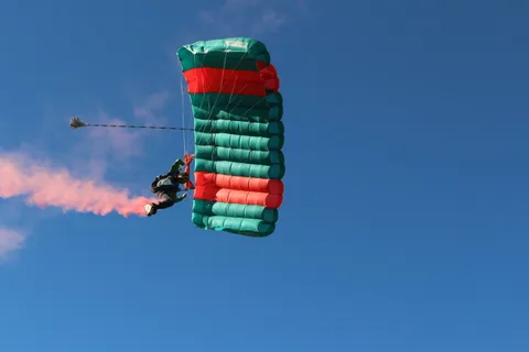 A skydiver glides under a green and red canopy trailing pink smoke against a clear blue sky.
