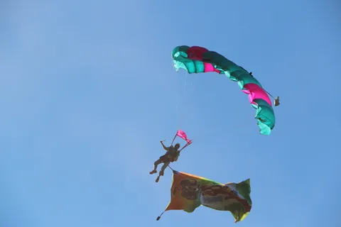 A skydiver carrying a flag banner falls free beneath a teal and pink parachute.