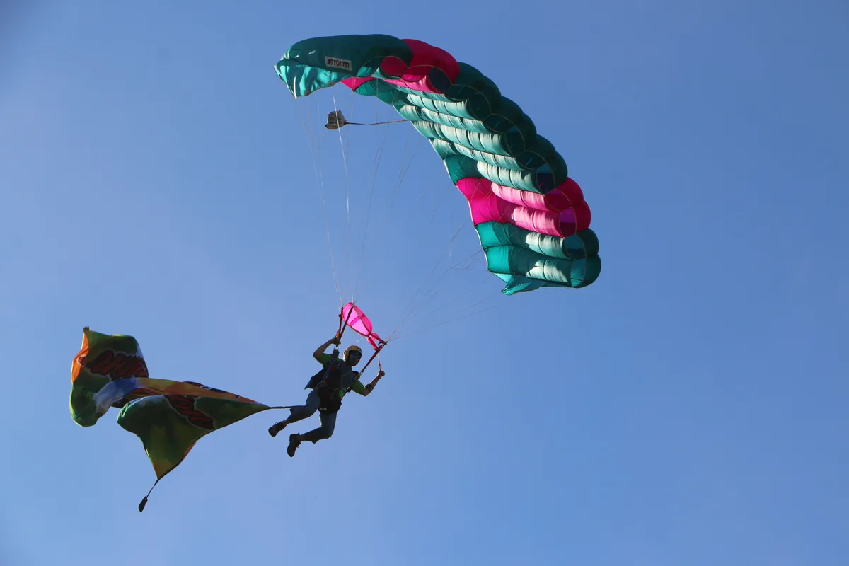 A skydiver holds a flag banner beneath a teal and pink parachute against a clear sky.